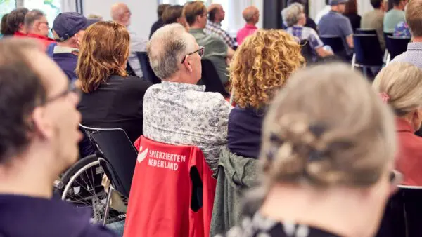 Veel personen luisteren aandachtig naar een lezing tijdens een bijeenkomst van Spierziekten Nederland.
